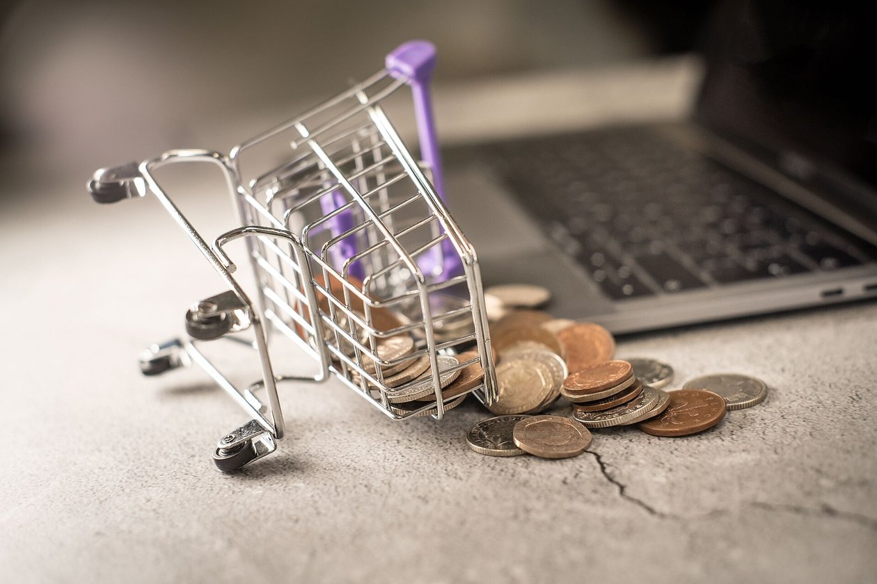A small metal shopping cart lying on its side with coins spilled beside it, an open laptop keyboard in the background